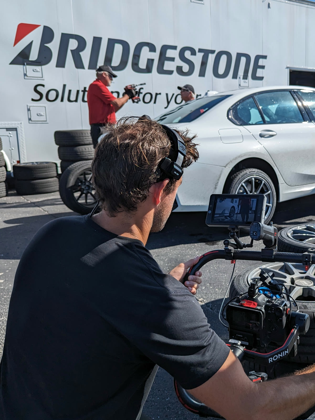 Close on the rig at a Bridgestone track day: an operator's gimbal and monitor in the foreground, stacked tyres and a white test sedan behind, the branded transporter filling the background