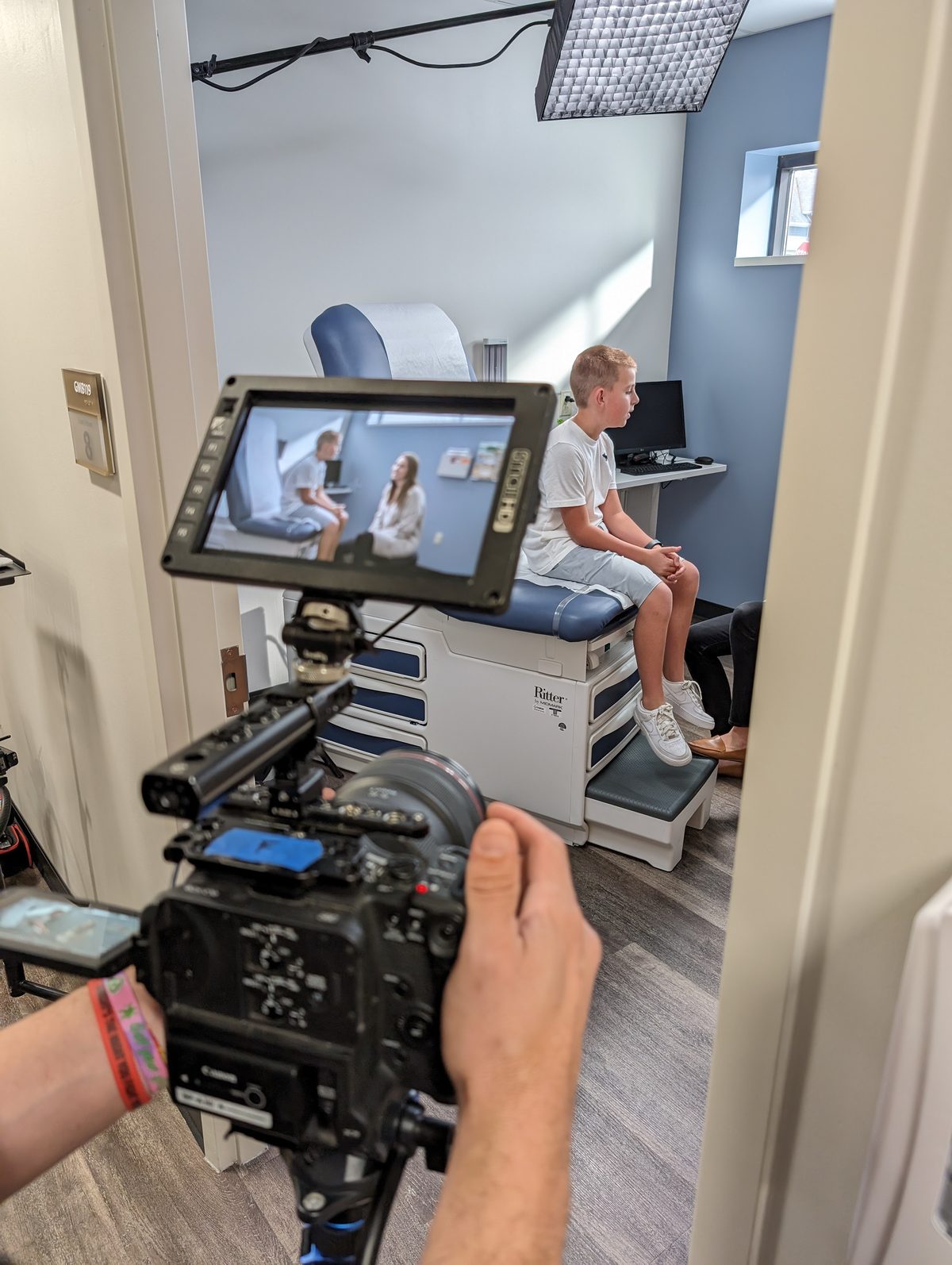 A Tactiv operator frames a young patient on an exam table through a cinema camera, the director's monitor showing the clinician and patient in shot