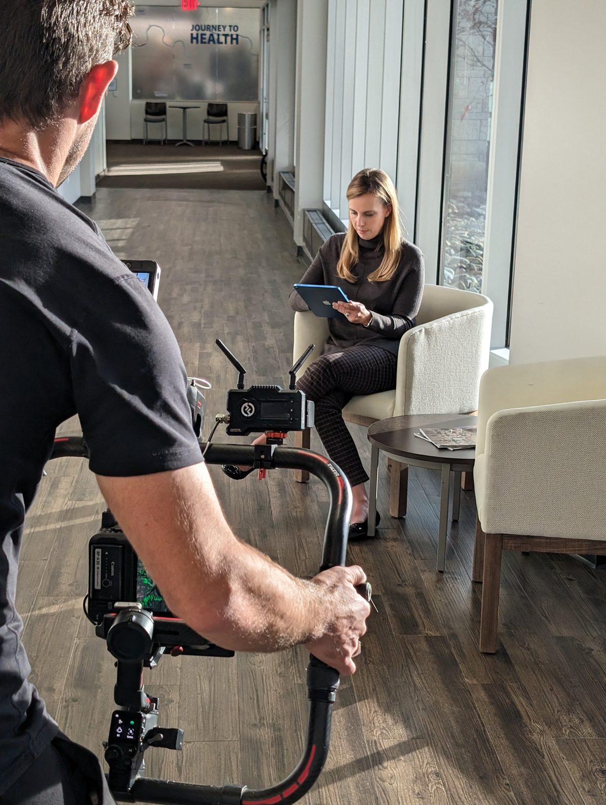 A Tactiv operator tracks a woman working on a tablet in a bright clinic lobby with a gimbal-mounted cinema camera