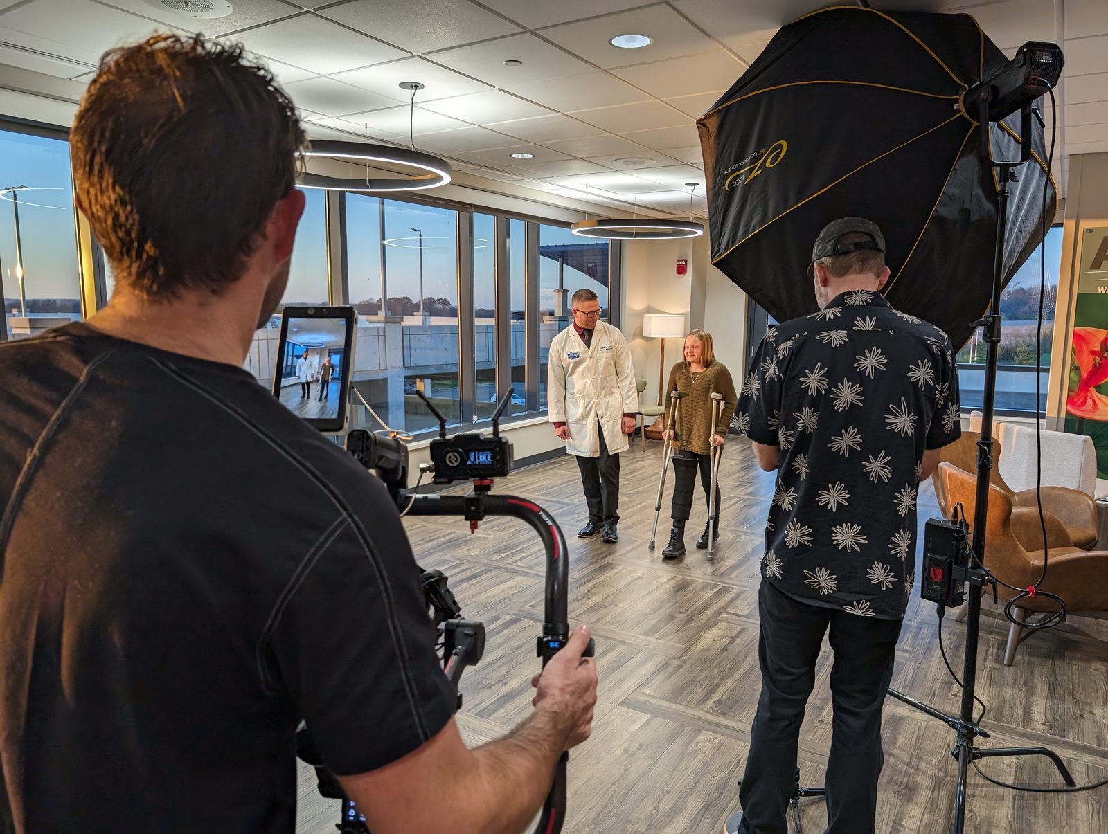 Tactiv crew filming a physician walking beside a patient on crutches in a hospital rehabilitation lobby, lit by a large octabox