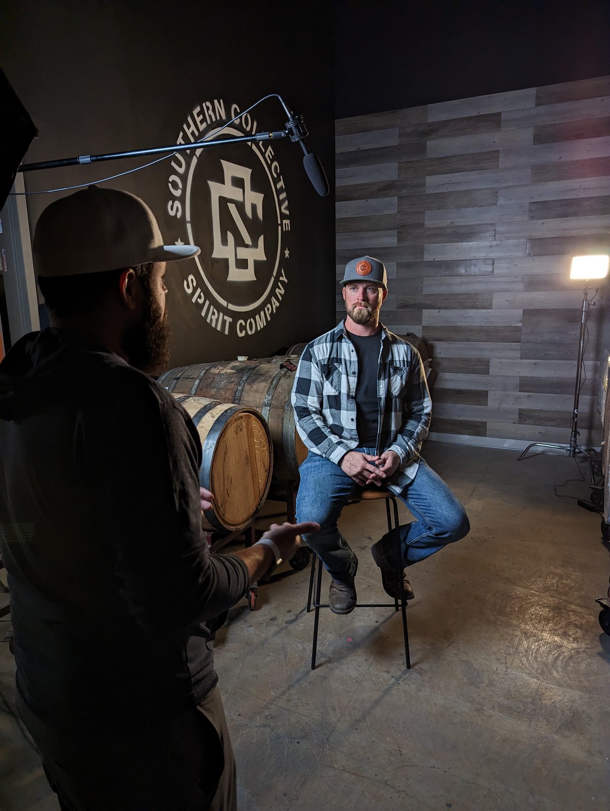 A Tactiv director cues a bearded subject seated in front of a distillery brand wall and a bourbon barrel, boom microphone extended overhead