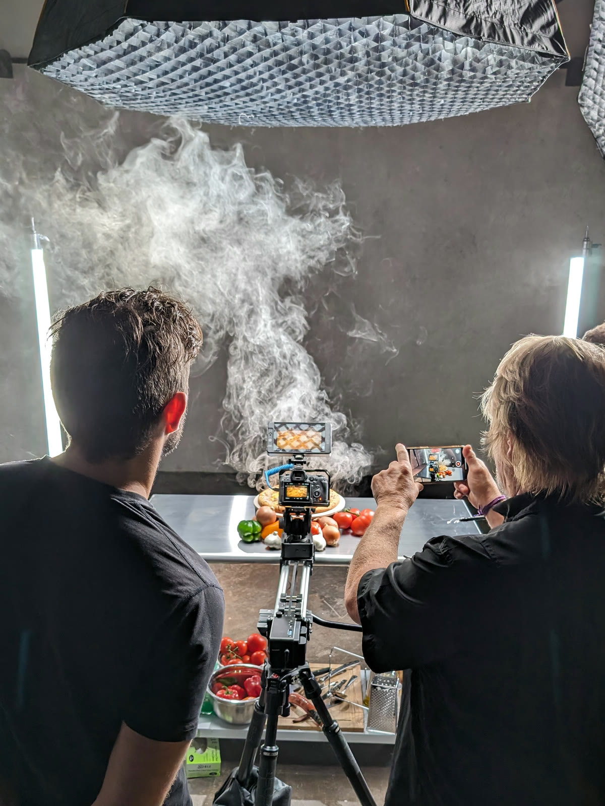 Two Tactiv crew watch a tabletop food build come together: a camera locked off on a slider, strip lights either side, and a plume of haze rising behind the dish