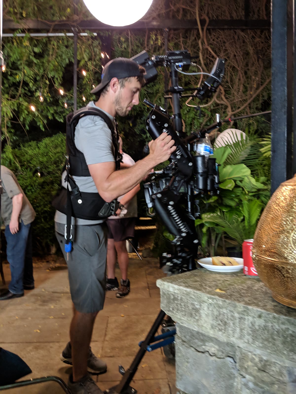 A Tactiv operator balances a cinema camera on a stabiliser vest and arm at an evening shoot, string lights and planting behind him and a plated dish on the stone counter in front