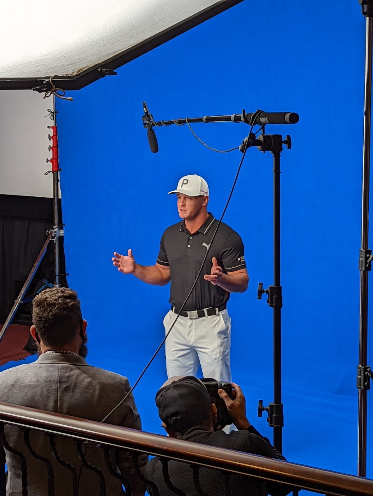 Bryson DeChambeau delivers a piece to camera on a blue-screen stage during a Bridgestone Golf shoot, boom microphone overhead and the client watching from the gallery rail