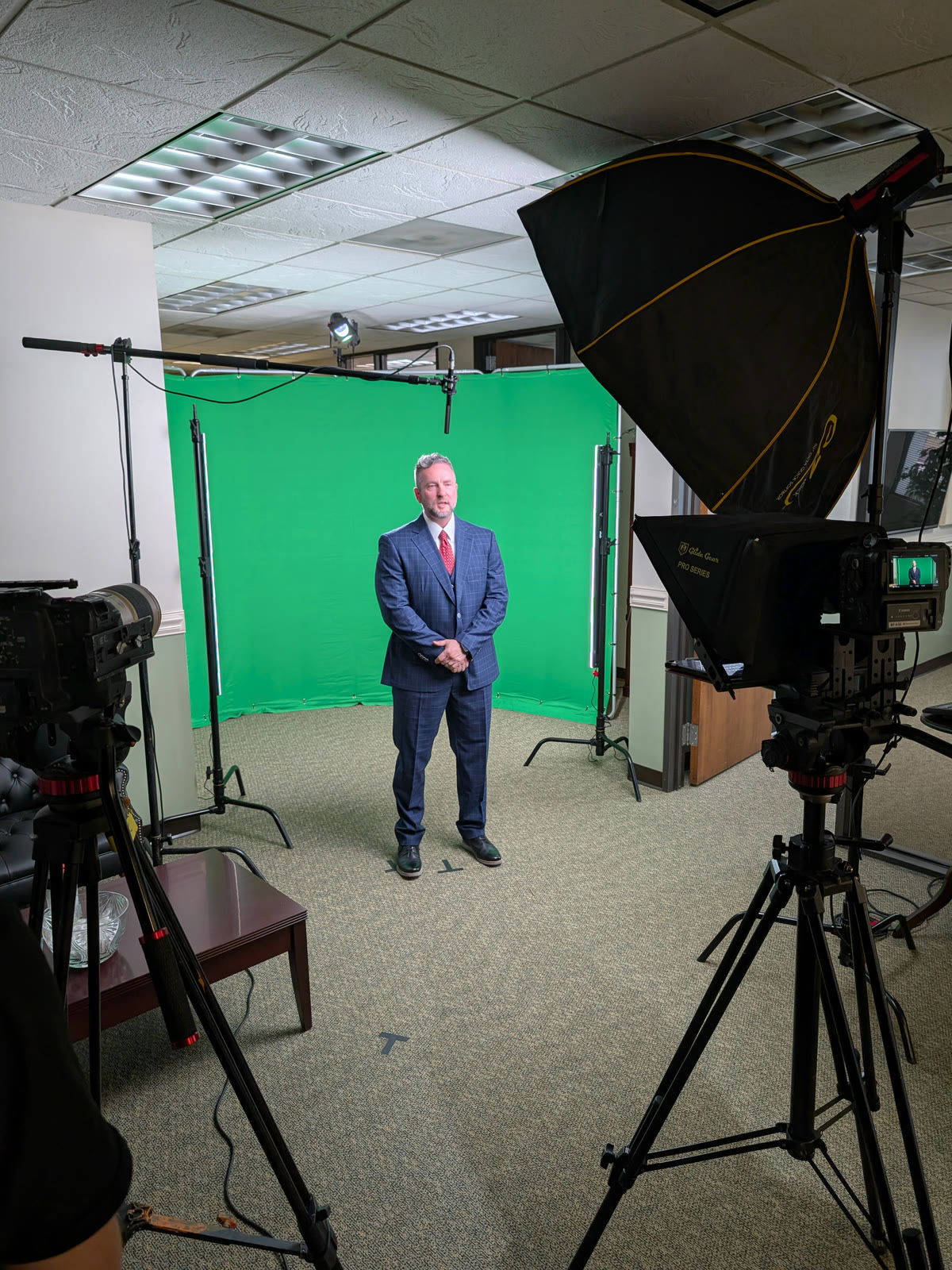 A green-screen build seen from the lighting side: the spokesman on his mark, a large octabox feathered in from camera right and a prompter-mounted camera looking back at him