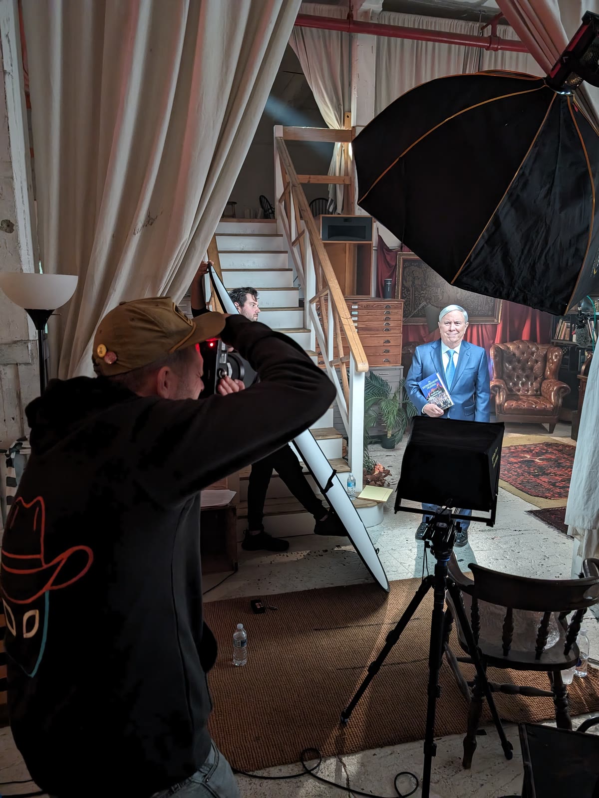 A stills photographer shoots a portrait set built inside a house: subject on his mark against a staircase and rug, a large octabox rigged overhead and a tethered monitor beside the tripod