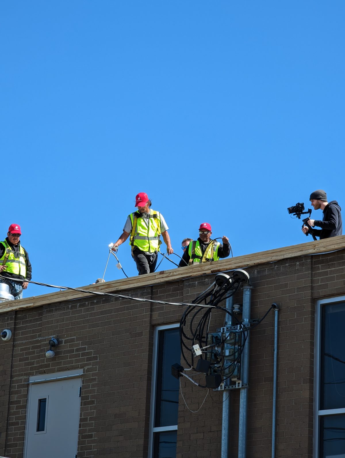 A Tactiv operator on a commercial rooftop films a crew in hard hats and hi-vis vests working a cable run against a clear blue sky
