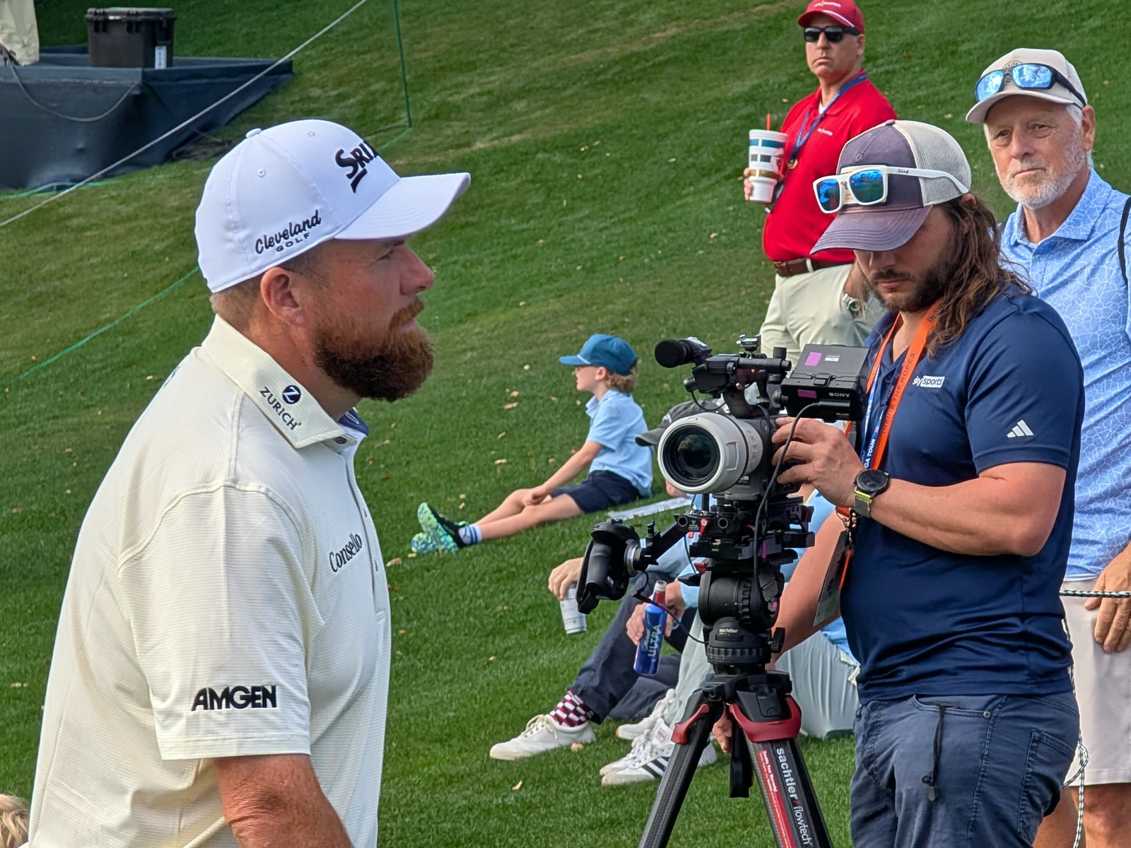 Inside the ropes on a tournament day: a Tactiv operator works a tripod-mounted cinema camera a few feet from a tour professional walking off the green, the gallery banked behind them