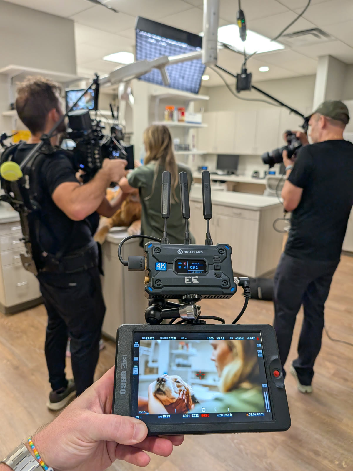 A director's monitor held in frame on a veterinary shoot, showing a dog on the treatment table while an operator on a support vest and a second camera work the room behind it