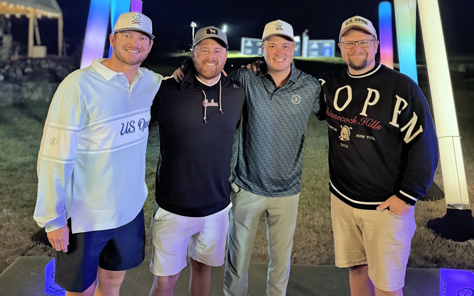 Four men in U.S. Open and golf-brand apparel stand arm in arm at a night event beside a floodlit course, colour-lit inflatable columns and a lit hospitality tent behind them