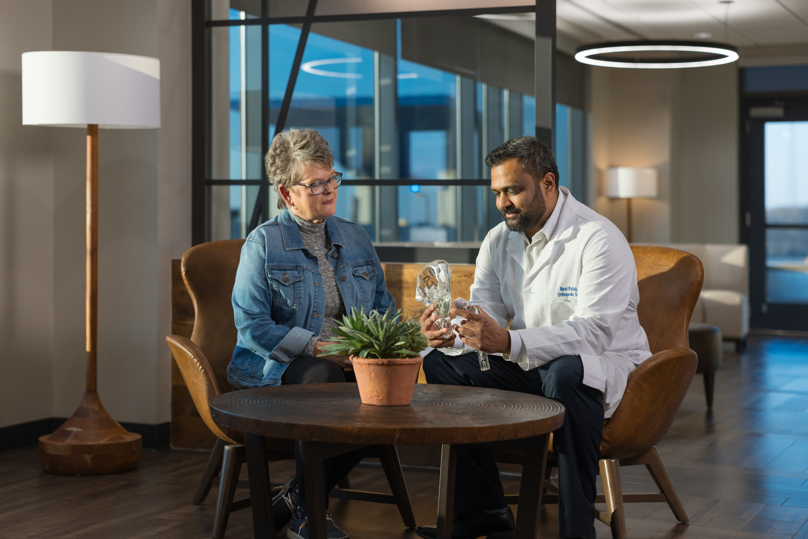 ADENA Health campaign still: a clinician shows an anatomical joint model to a seated patient across a low table in a consultation lounge