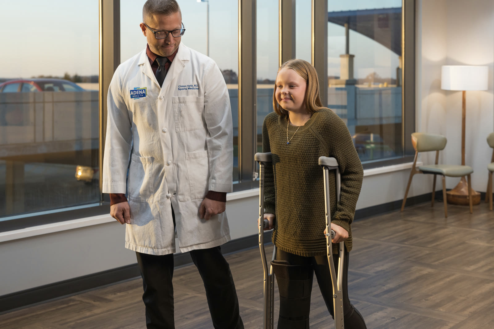 ADENA Health campaign still: a physician in a branded white coat walks a young patient on crutches through a daylit hospital lounge