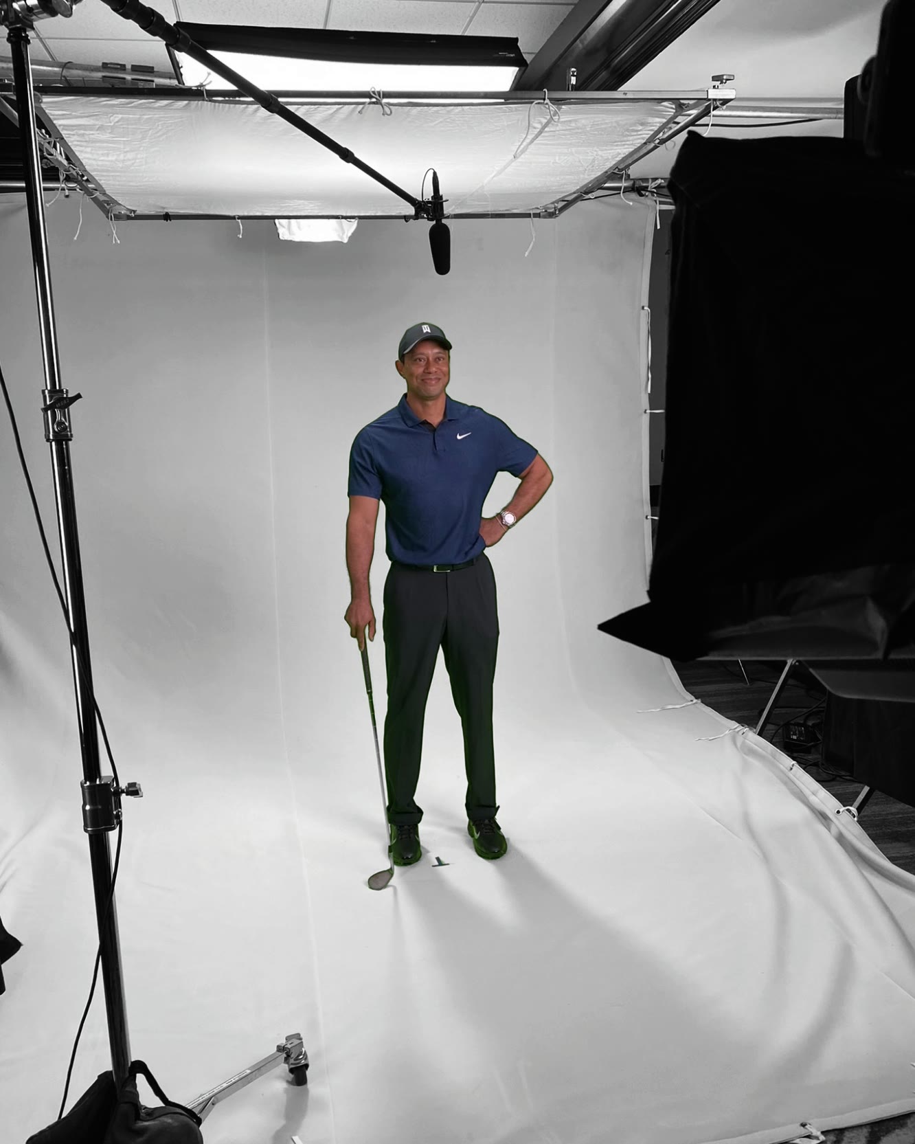 Tiger Woods on a white cyclorama set during a studio production: an overhead diffusion frame and boom microphone dropped in from the top of the set, black flags and C-stands to either side