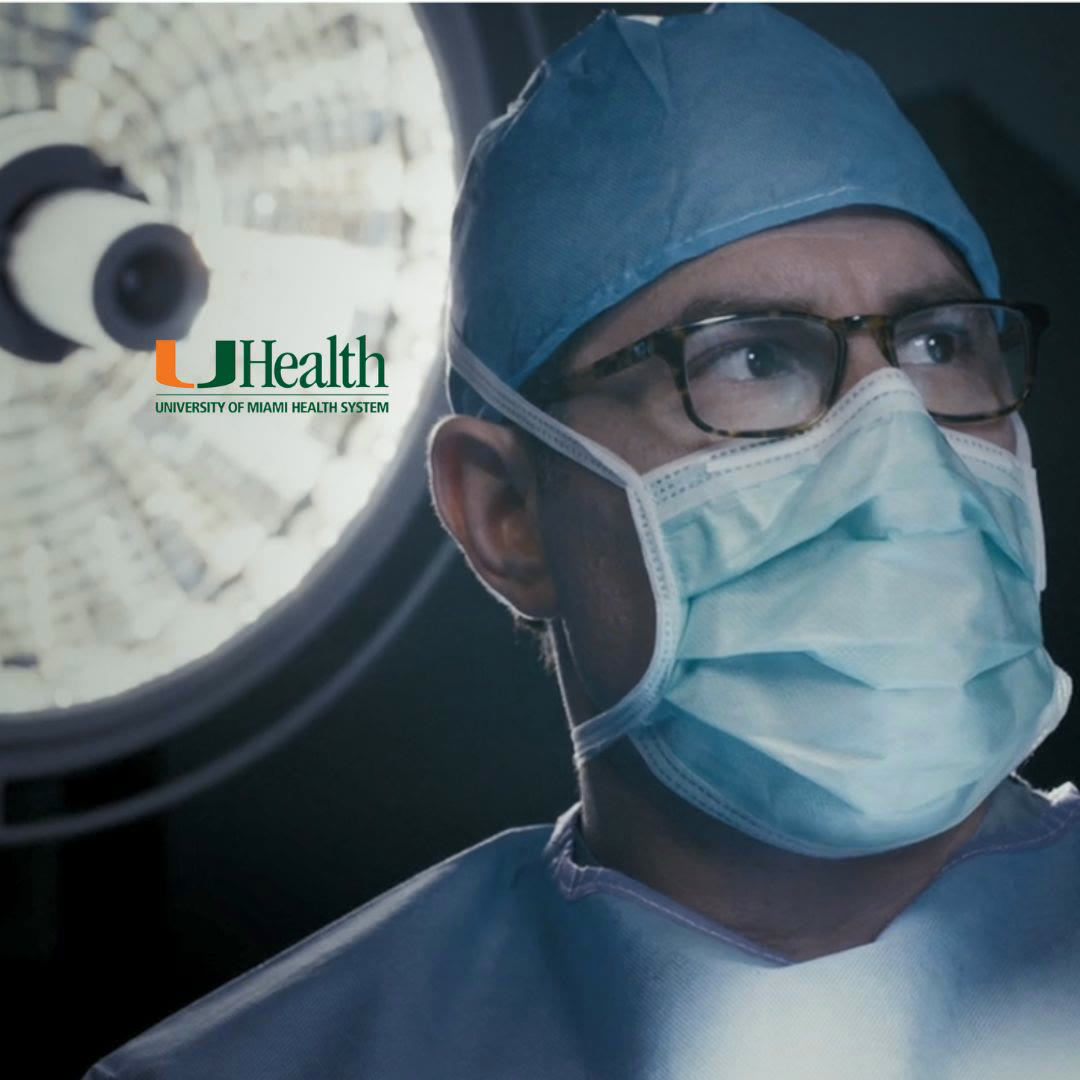 Frame from a UHealth brand film: a masked surgeon looks off camera under an operating-theatre light, the University of Miami Health System mark set into the frame